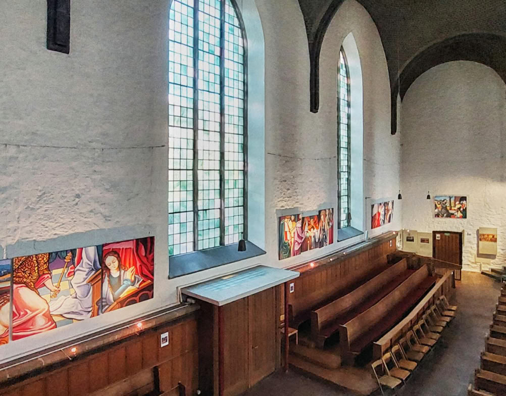 Das Bild zeigt den Innenraum der Altstädter Nikolaikirche in Bielefeld. Die Wände sind weiß verputzt, während die hohen Gewölbedecken aus dunklem Mauerwerk bestehen. An den Seitenwänden befinden sich große, schlanke Rundbogenfenster, die mit vielen kleinen, klaren Glasquadraten (Sprossenfenster) ausgestattet sind und viel Tageslicht hereinlassen.  Unterhalb der Fenster ziehen sich moderne, bunte Wandbilder entlang. Sie zeigen verschiedene Personen in lebhaften Farben (vor allem Rot, Orange, Blau und Weiß) und wirken wie Fresken oder großformatige Gemälde, die die biblische Geschichte oder Heilige darstellen.  Im unteren Bereich des Raumes stehen traditionelle Holz-Kirchenbänke. Sie sind auf der linken Seite in einer gestaffelten Anordnung und auf der rechten Seite in geraden Reihen aufgestellt. Der Boden ist hell und scheint aus Stein oder Beton zu sein. Im hinteren Bereich des Bildes ist eine Holztür sichtbar, und an der rückwärtigen Wand hängen weitere kleine gerahmte Bilder.